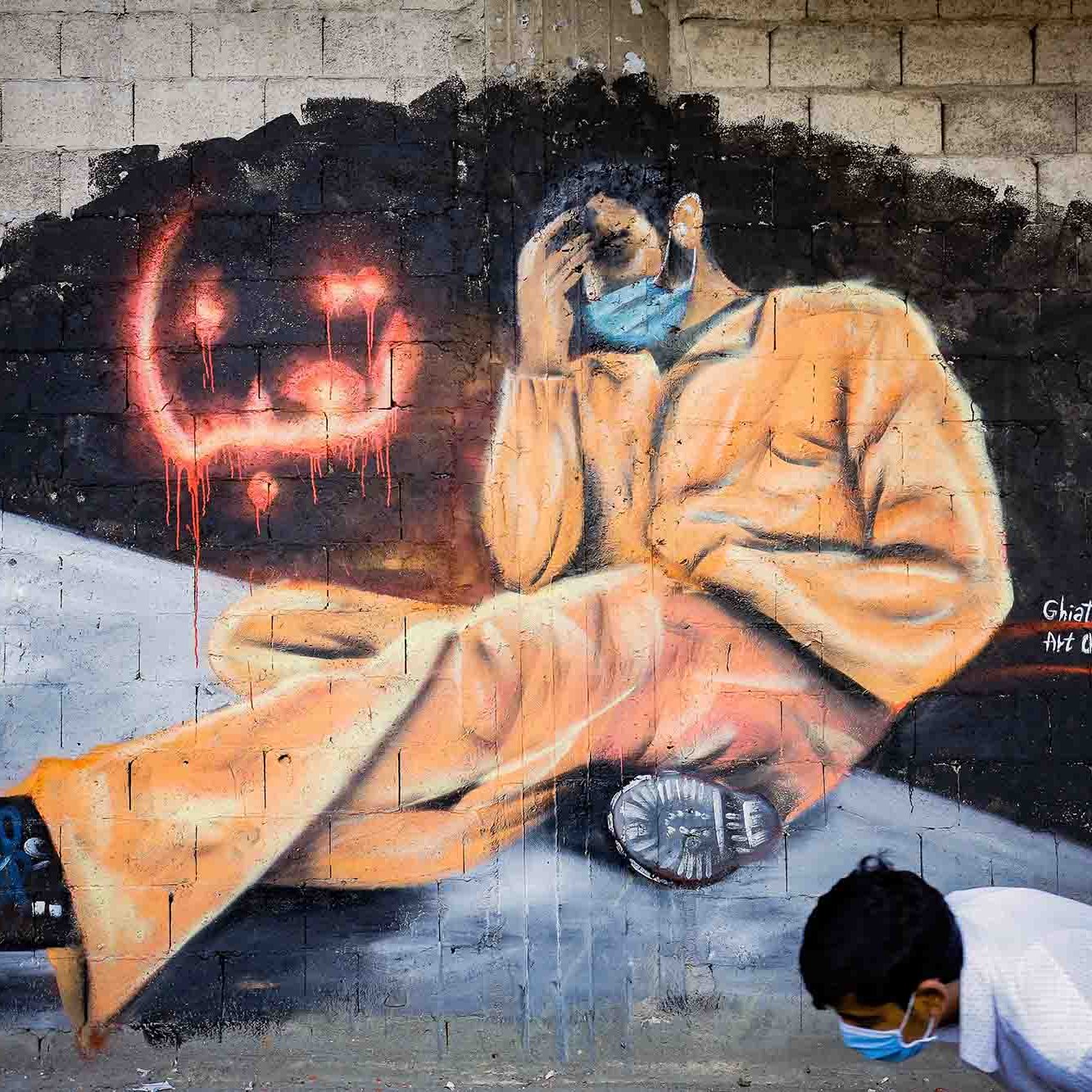 L'image représente une fresque murale colorée montrant un homme assis, habillé d'une combinaison orange. Il semble pensif, la main sur le visage, dans une posture qui évoque la détresse ou la réflexion. Au fond, on aperçoit des graffiti en rouge qui ajoutent une ambiance dramatique à l'œuvre. Un enfant, portant un masque, est également visible en bas de l'image, probablement en train d'observer ou d'interagir avec la fresque. L'ensemble transmet un message puissant lié à des thèmes de changement ou de lutte.
