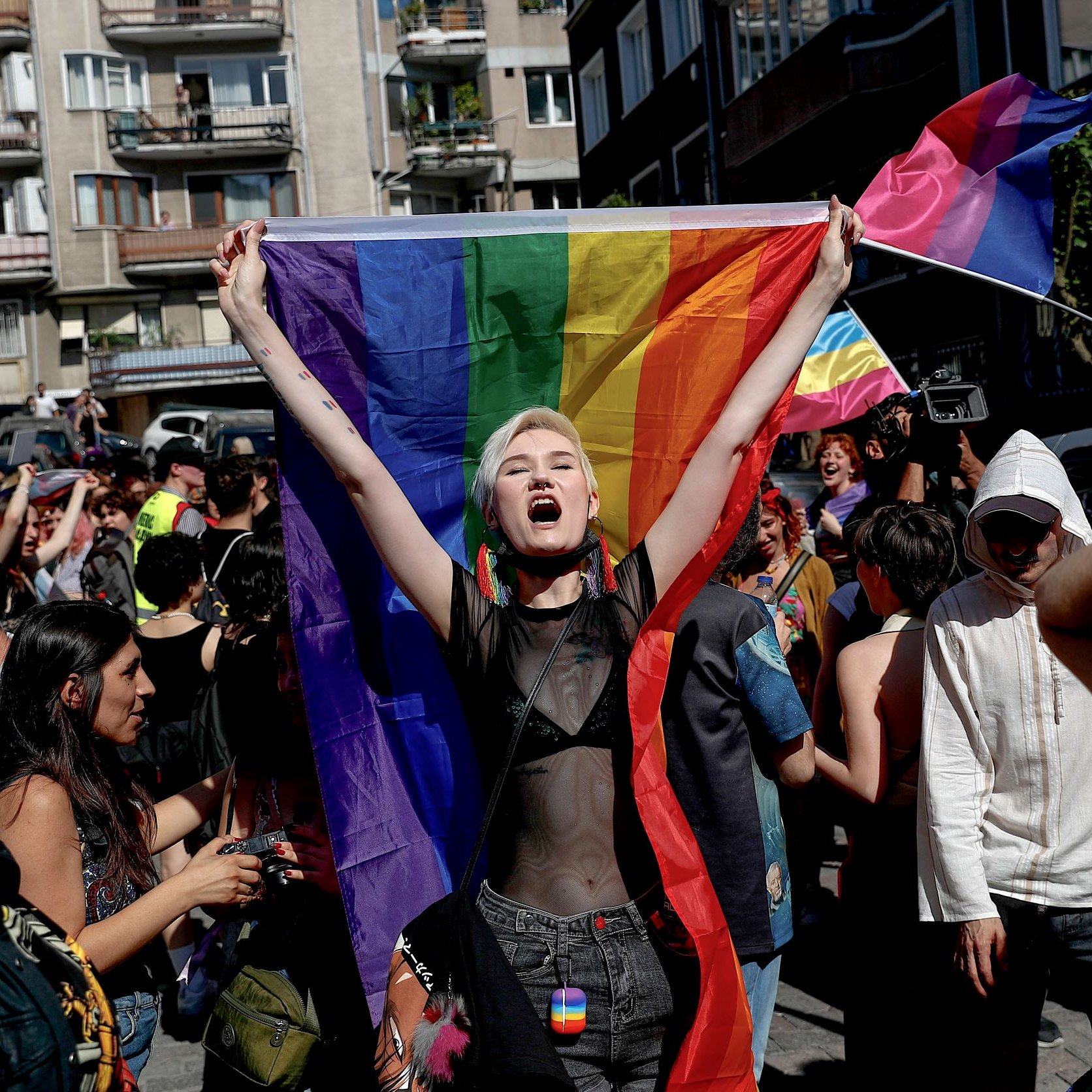 L'immagine mostra un gruppo di persone in una strada affollata, probabilmente durante una manifestazione o una parata. Al centro, una giovane donna con capelli corti e bianchi è in primo piano, mentre tiene alta una grande bandiera arcobaleno. Sembra esprimere gioia e determinazione. Intorno a lei, altre persone sventolano bandiere e manifestano il loro sostegno, creando un'atmosfera di festeggiamento e orgoglio. Le decorazioni e i colori vivaci trasmettono un messaggio di inclusione e solidarietà.