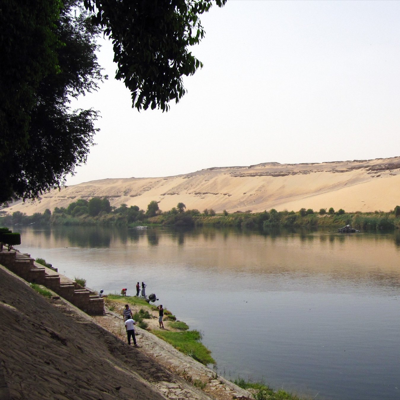 L'image montre un paysage paisible où un fleuve serpente le long d'une rive. Sur un côté, il y a des dunes de sable doré qui montent en pente, créant un contraste avec la verdure des arbres qui bordent le fleuve. Des personnes se trouvent au bord de l'eau, peut-être en train de pêcher ou de profiter de la nature. Le ciel est clair, et la lumière dubaine donne une atmosphère sereine à la scène.