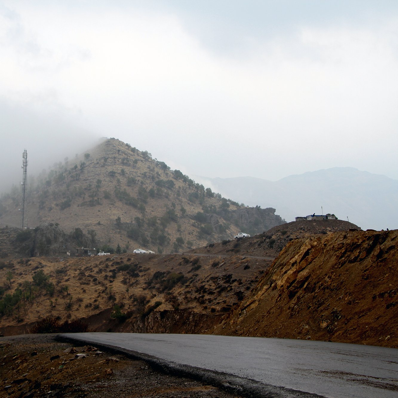 در این تصویر، یک جاده‌ا‌ی در کوهستان با شیب ملایم دیده می‌شود. در سمت راست، جاده به سمت دورتر ادامه پیدا می‌کند و در بالای تپه‌ها، ابرها در حال پایین آمدن و محو کردن قله‌ها هستند. روی تپه‌ها، درختان نسبتاً کوچک و فضاهای خشک دیده می‌شوند. در بالای تپه، یک دکل تلفن همراه نیز به چشم می‌خورد و چندین خودروی سفید رنگ در دور دست قابل مشاهده‌اند. جو تصویر ابری و مرطوب به نظر می‌رسد.