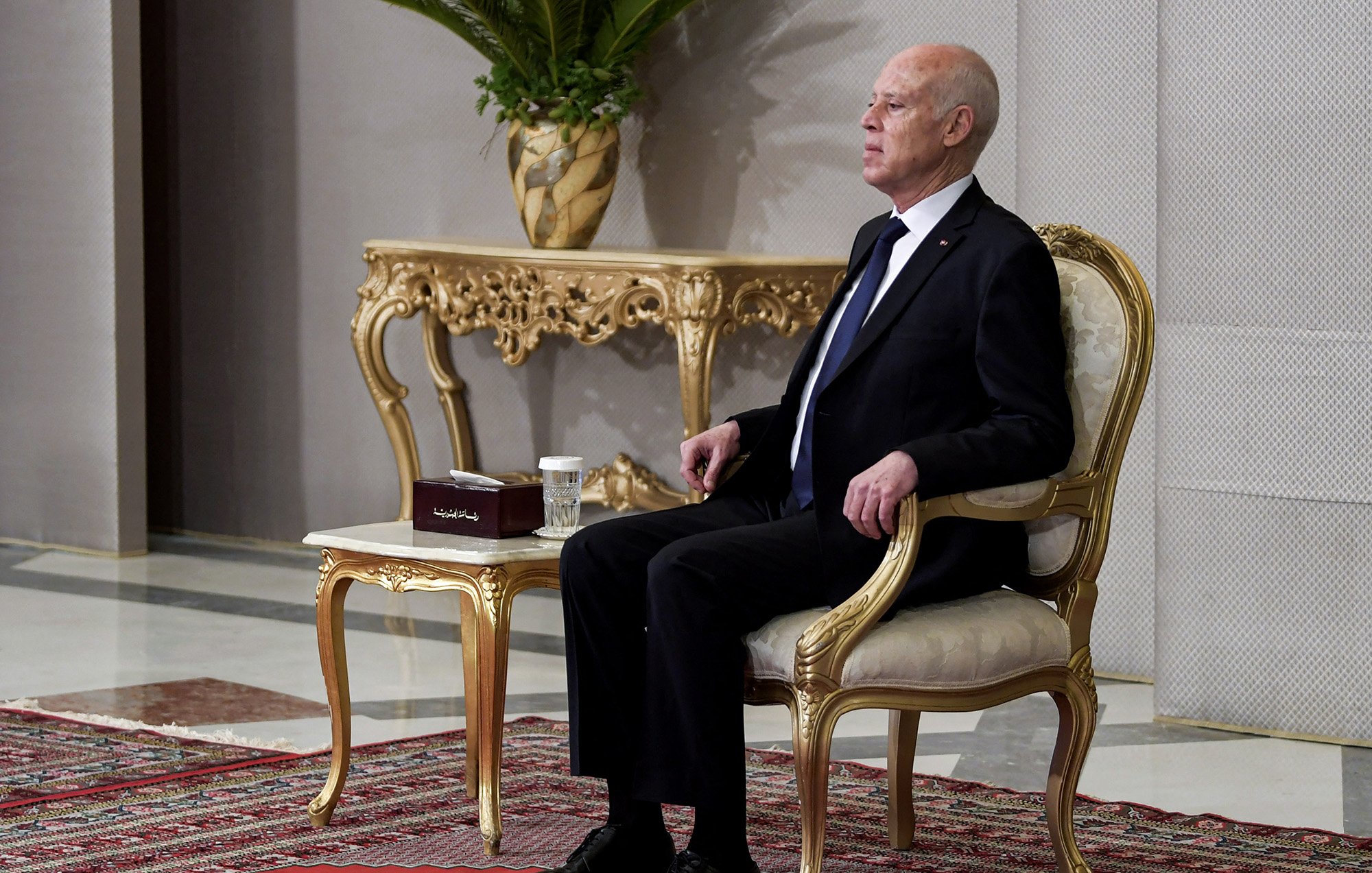 L'image montre un homme assis sur une chaise élégante dans un cadre formel. Il porte un costume noir avec une cravate. À côté de lui se trouve une table ornée sur laquelle repose une petite boîte et une tasse. L'arrière-plan présente des murs décorés et une plante en pot, ajoutant une touche de sophistication à l'environnement. Le sol est recouvert d'un tapis, contribuant à l'atmosphère formelle de la scène.