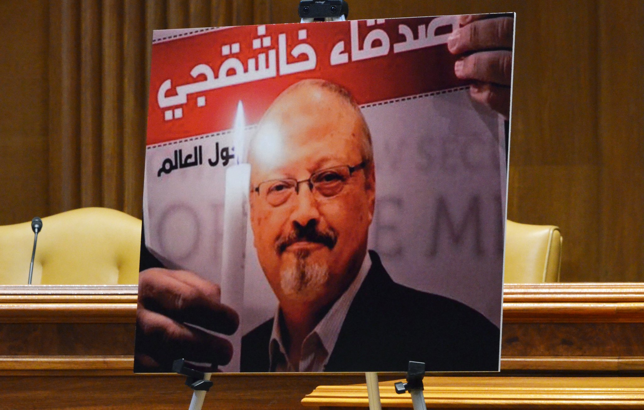 L'image montre un portrait d'un homme tenant une bougie, accompagné d'un texte en arabe et en français. Le fond de l'affiche évoque un hommage ou un soutien, probablement lié à une question de justice. Le contexte global semble être une commémoration ou une manifestation pour défendre les droits humains. Deux chaises sont visibles en arrière-plan, suggérant que l'affiche est présentée lors d'un événement formel.