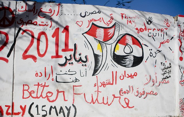 The image features a wall covered in graffiti and artwork. Prominent elements include the Egyptian flag, the date "25 January 2011," and references to the revolution. Phrases in Arabic and English express themes of hope, such as "Better Future" and "إرادة," meaning "will" or "determination." The background suggests a lively and politically charged atmosphere, symbolizing the spirit of change during the Egyptian revolution. Various colors like red, black, and white are used, enhancing the visual impact of the message.