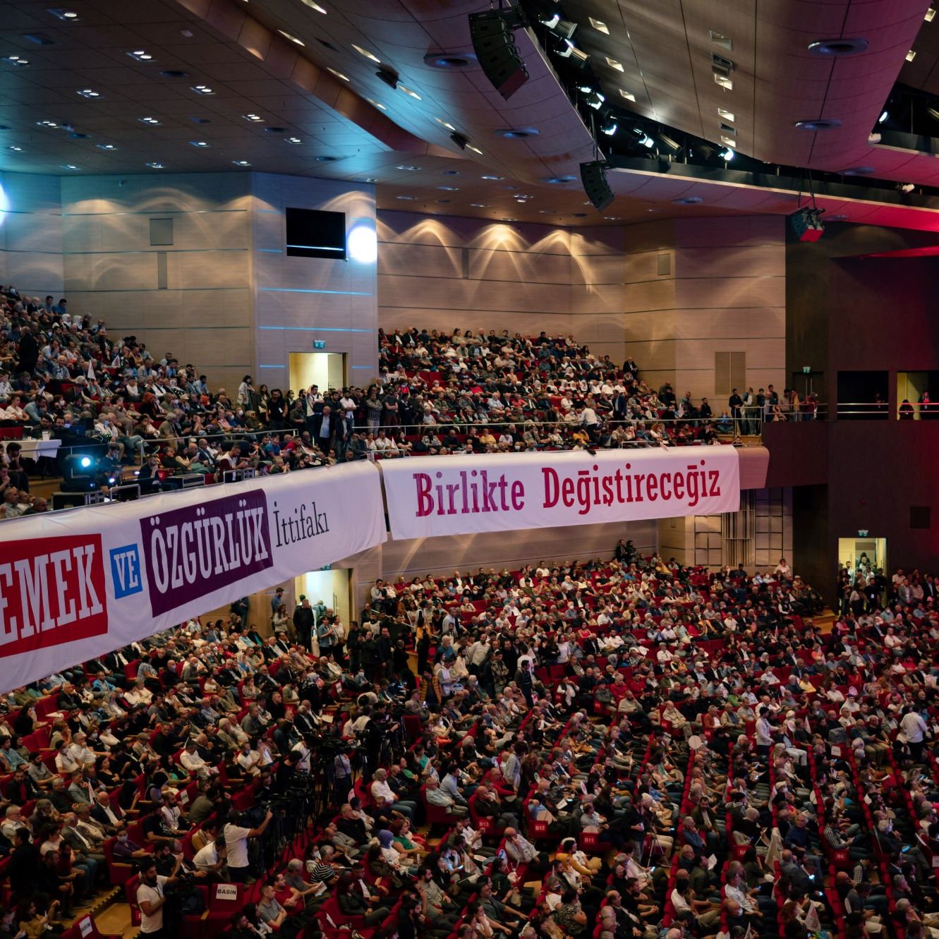 در این تصویر، صحنه‌ی یک سالن بزرگ کنفرانس با تعدادی بسیار زیاد از افراد در حال نشستن و تماشا دیده می‌شود. سالن به خوبی روشن است و روی دیوارهای آن نوشته‌هایی به زبان ترکی وجود دارد. یکی از این نوشته‌ها به صورت برجسته "EMEK VE ÖZGÜRLÜK" (به معنی "کار و آزادی") است. فضا به نظر می‌رسد مربوط به یک همایش یا تجمع سیاسی باشد که در آن مردم به موضوعاتی مهم توجه دارند. در پس‌زمینه، نورپردازی خاصی وجود دارد که بر احساسات و فضای کنفرانس تأکید می‌کند.
