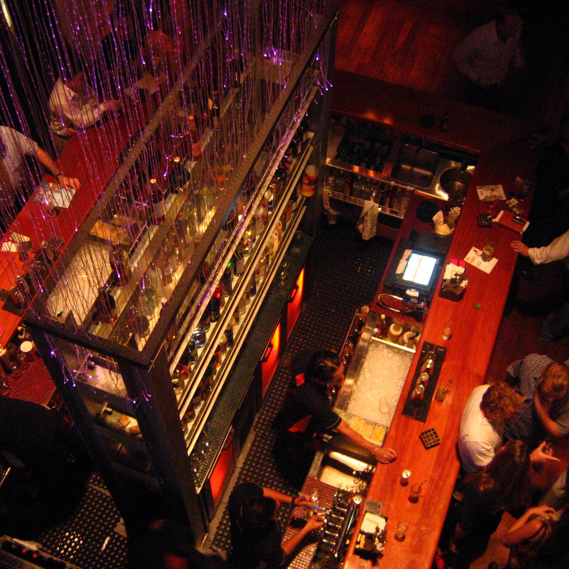 L'image montre une vue aérienne d'un bar animé. On peut voir une grande table de bar en bois clair, avec de nombreuses personnes stationnées autour, se parlant et déguster des boissons. Au dessus, il y a des chaînes de lumières, créant une ambiance festive. Les étagères à bar sont bien garnies de bouteilles colorées, et on distingue des serveurs s'affairant derrière le comptoir. L'atmosphère semble conviviale et dynamique, avec une décoration moderne.
