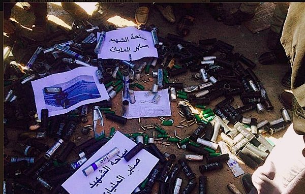 L'image montre un grand nombre de cartouches de munitions et de projectiles disposés au sol. Certaines de ces munitions sont accompagnées de panneaux ou de feuilles de papier avec des inscriptions en arabe. On peut voir des personnes autour de cette disposition, mais leurs visages ne sont pas visibles. L'ensemble de la scène semble évoquer une situation liée à la confiscation ou à la destruction d'armements.