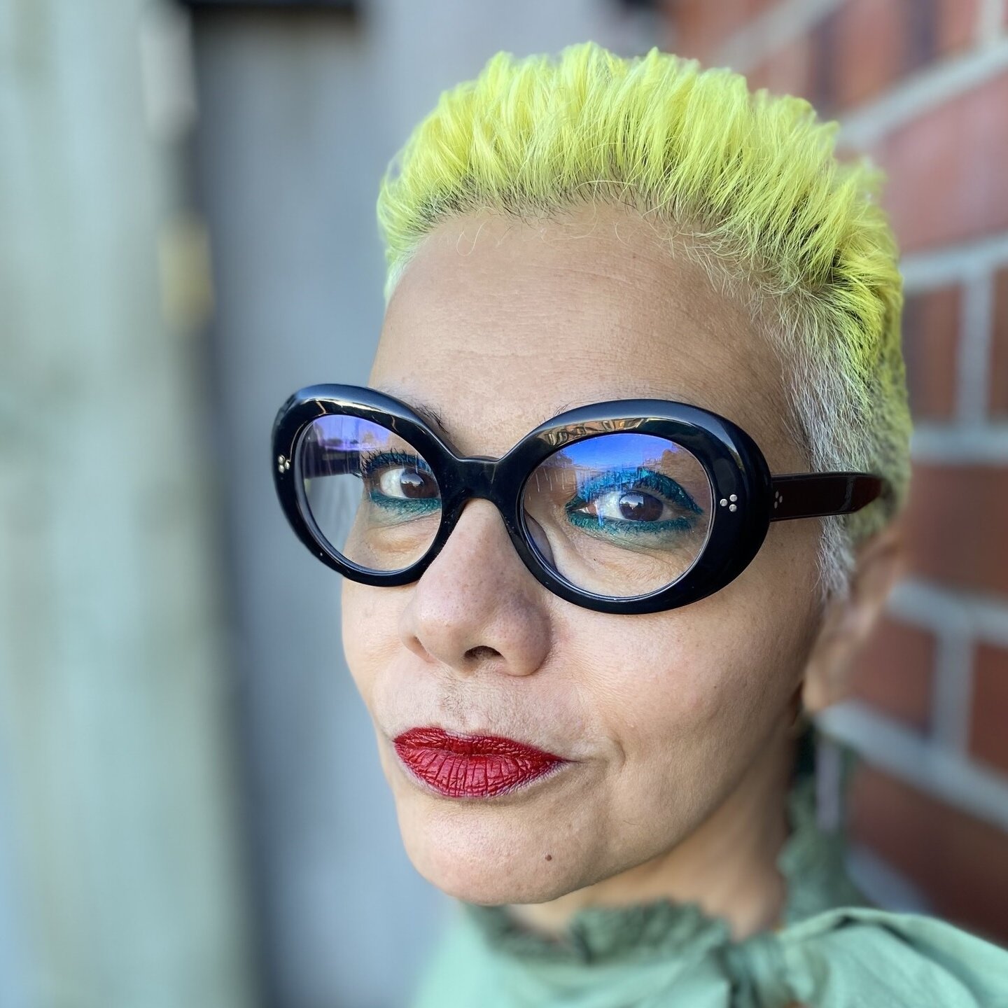 L'image montre une personne avec des cheveux très courts teints en jaune vif. Elle porte de grandes lunettes noires au style distinctif. Son maquillage comprend des lèvres rouge vif. Le fond présente un mur en briques, ce qui ajoute un contraste avec son look. Elle a une expression confiante et souriante.