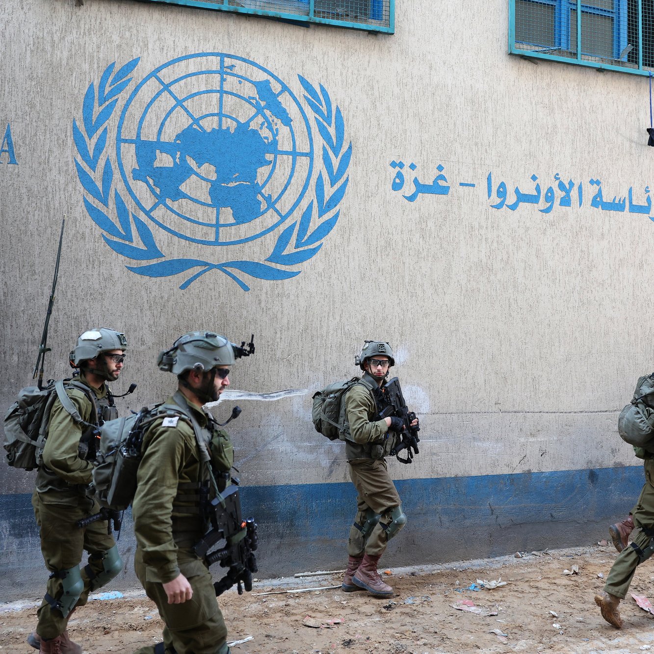 در این تصویر، تعدادی نیروهای نظامی در حال حرکت در کنار یک ساختمان به چشم می‌خورند. دیوار ساختمان با نوشته‌ای به زبان عربی و علامت سازمان ملل تزئین شده است. این نیروها لباس‌های نظامی و تجهیزات مناسب به تن دارند. به نظر می‌رسد که مکان مربوط به نهادهای بین‌المللی مانند یونروا (UNRWA) در غزه باشد. فضای اطراف دارای حالت فعالیت و حرکت است.