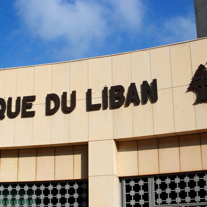 L'image montre un bâtiment avec l'inscription "BANQUE DU LIBAN" en lettres majuscules. La façade est faite de pierre ou de béton, et il y a un symbole d'un petit arbre à côté du texte. Le ciel en arrière-plan est bleu avec quelques nuages. L'ensemble donne une impression de modernité et de solidité.