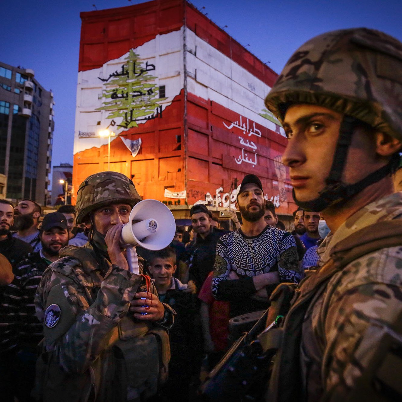 L'image montre un groupe de personnes rassemblées, dont plusieurs soldats en uniforme. L'un des soldats utilise un mégaphone, probablement pour communiquer avec la foule. En arrière-plan, on peut voir un bâtiment peint avec les couleurs du drapeau libanais. L'atmosphère semble tendue et engagée, suggérant une situation de manifestation ou de rassemblement important. La lumière est douce, indiquant peut-être le crépuscule. Les expressions des participants sont variées, allant de la concentration à l'écoute attentive.