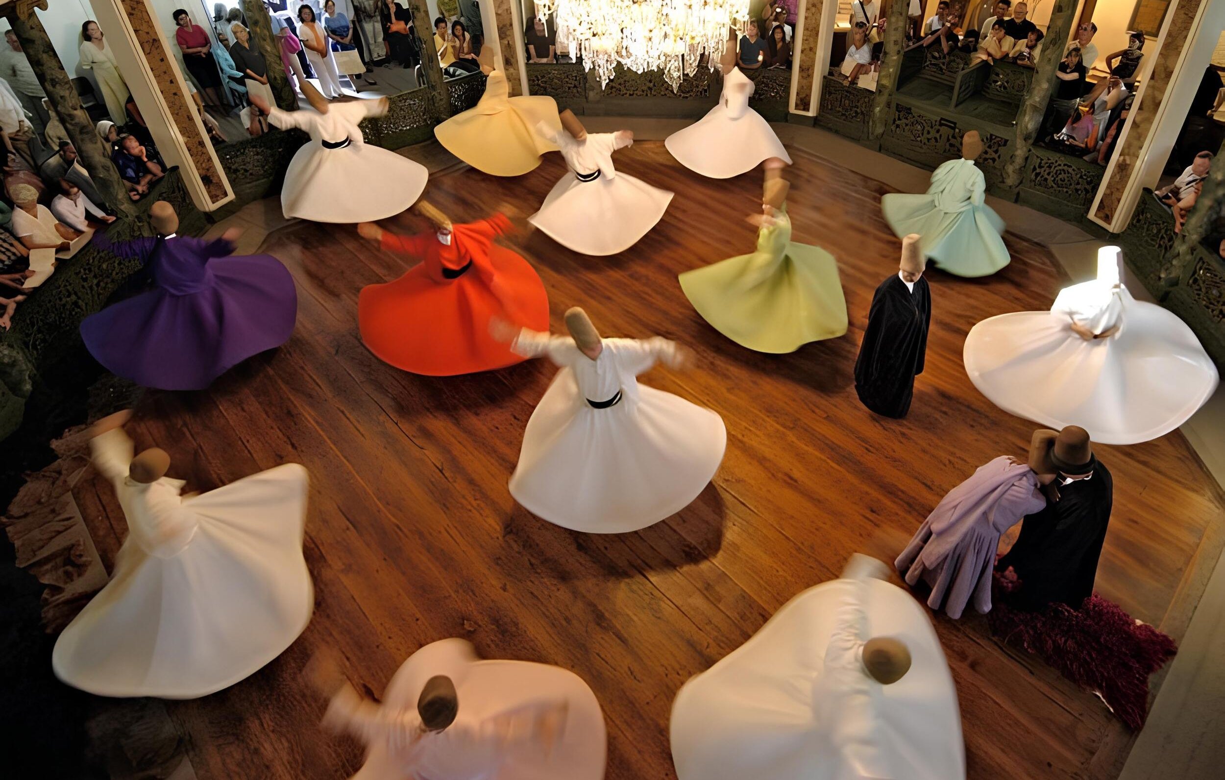 L'immagine mostra un gruppo di ballerini che si esibiscono in una danza tradizionale, con abiti ampi e colorati che girano in cerchio. I ballerini sono disposti in un grande spazio circolare, con un pavimento in legno lucido e una decorazione elegante attorno a loro. Al centro della scena, un lampadario di cristallo illumina l'ambiente, mentre il pubblico osserva l'esibizione con interesse. I colori degli abiti, che vanno dal bianco al viola e arancio, creano un'atmosfera vibrante e festosa.