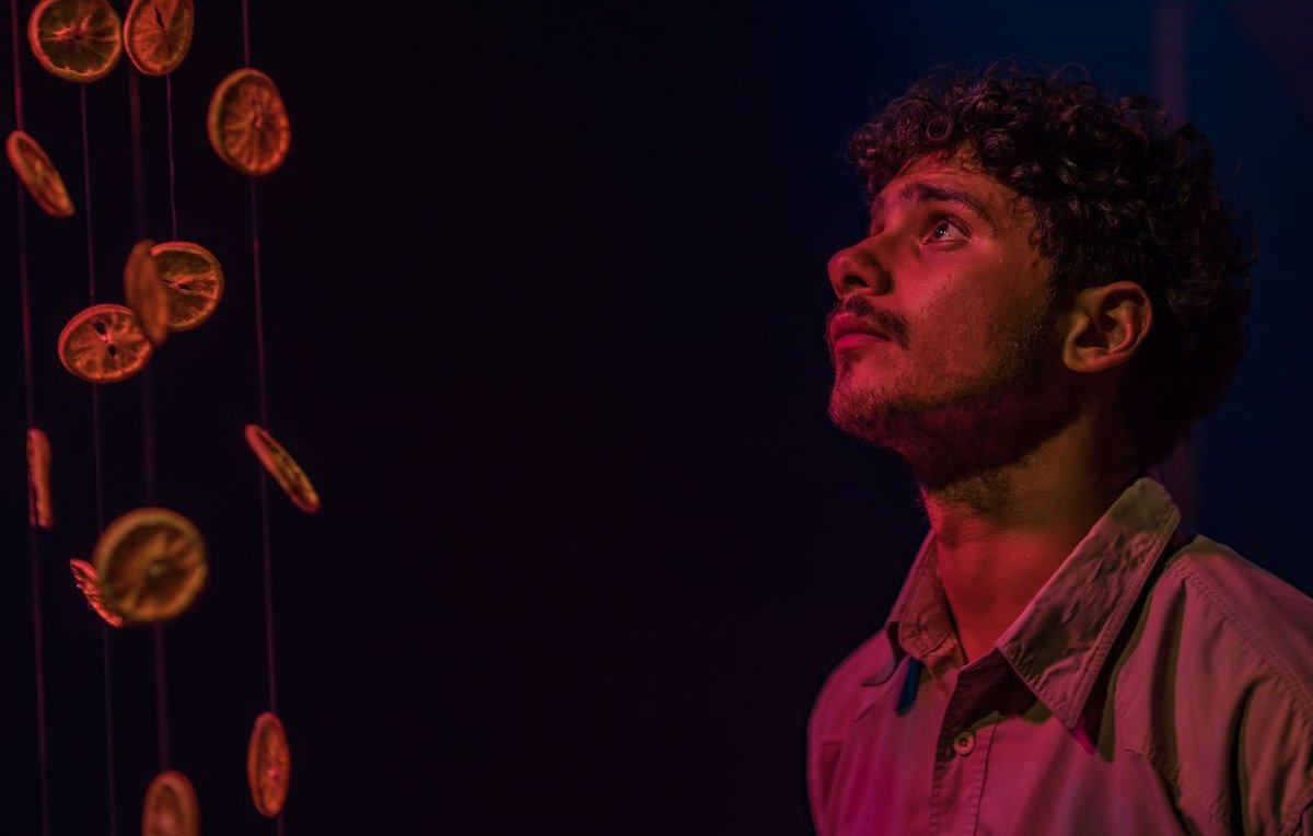L'image montre un jeune homme regardant avec curiosité quelque chose au-dessus de lui. Il est entouré d'une ambiance sombre, avec des éclairages qui créent des reflets colorés, probablement des nuances de bleu et de rose. Des rondelles d'orange sèchées pendent de fil, créant un effet visuel intéressant, tandis que l'expression du jeune homme semble pensive et captivée par la scène.