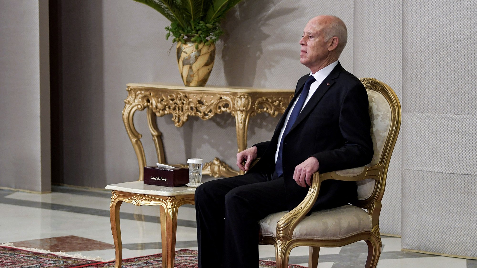 L'image montre un homme assis sur une chaise élégante dans un cadre formel. Il porte un costume noir avec une cravate. À côté de lui se trouve une table ornée sur laquelle repose une petite boîte et une tasse. L'arrière-plan présente des murs décorés et une plante en pot, ajoutant une touche de sophistication à l'environnement. Le sol est recouvert d'un tapis, contribuant à l'atmosphère formelle de la scène.