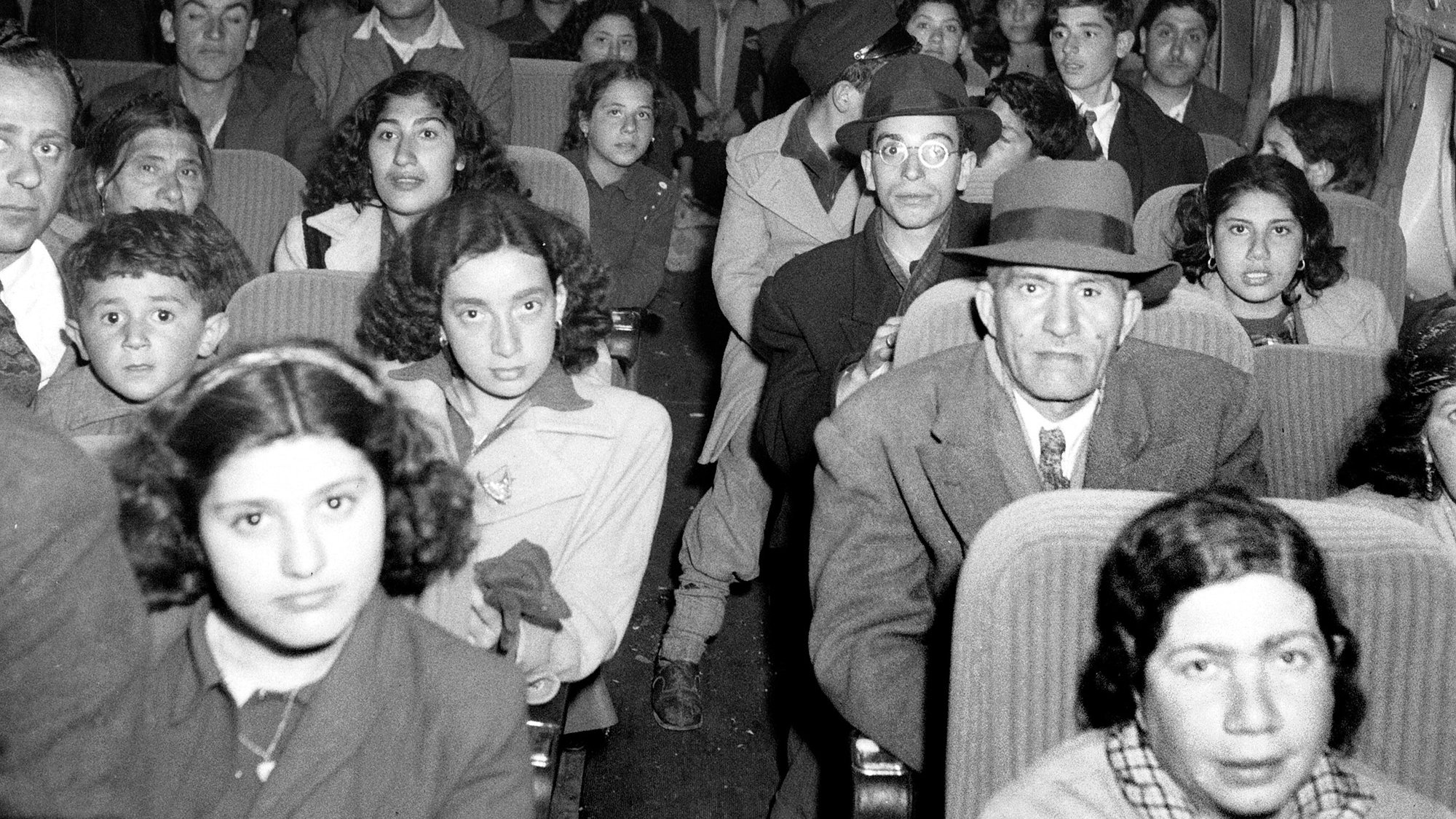 La imagen muestra el interior de un avión lleno de pasajeros. Hay muchos hombres, mujeres y niños sentados en los asientos con expresiones variadas. La mayoría de las personas parecen estar mirando hacia la cámara. El ambiente es de una época pasada, con un estilo de vestimenta que refleja un tiempo diferente. La iluminación es tenue, lo que contribuye a la atmósfera general de la fotografía.