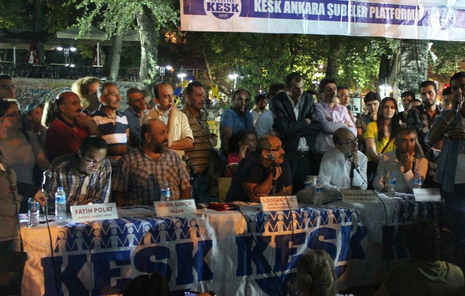 L'image montre une scène de rassemblement ou de meeting en extérieur. Une table est placée au premier plan, où plusieurs personnes sont assises, apparemment des intervenants, pendant qu'une autre personne, debout, s'adresse à l'audience. Un public se tient autour, écoutant attentivement. En arrière-plan, il y a une bannière avec des inscriptions, et l'ambiance semble être celle d'un événement communautaire ou d'une manifestation. Les participants semblent engagés dans une discussion animée.