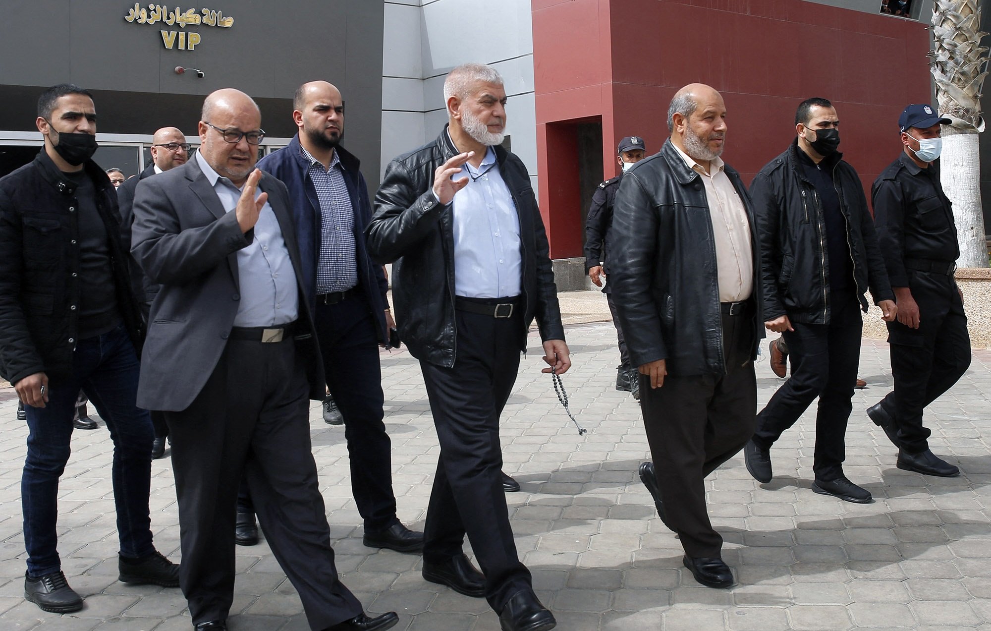 L'image montre un groupe de plusieurs hommes marchant ensemble à l'extérieur. Ils semblent se déplacer avec assurance et saluent peut-être des personnes présentes. Les hommes portent des vêtements variés, allant de vestes en cuir à des chemises et des pantalons sombres. L'environnement autour d'eux semble être un espace public, peut-être à proximité d'un bâtiment. Certains membres du groupe portent des masques. L'atmosphère paraît informelle mais organisée.