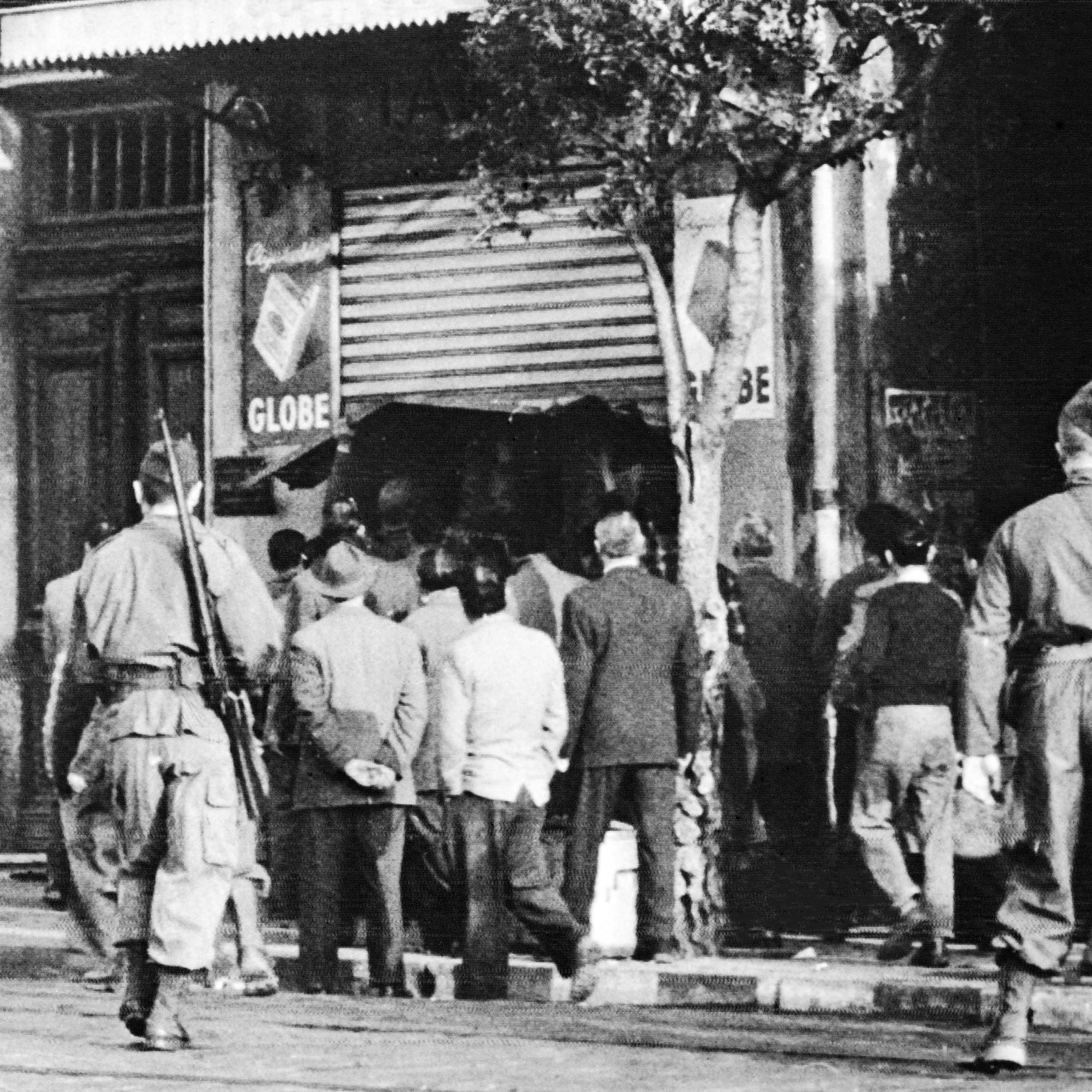 L'image montre une scène en noir et blanc où plusieurs soldats, en uniforme, se tiennent en arrière-plan. Devant eux, un groupe d'hommes en civil semble observer une situation à l'entrée d'un magasin ou d'un bâtiment, probablement une affaire en cours. La présence des soldats suggère un contexte de tension, peut-être lié à des événements historiques. Les détails de l'environnement, tels que les enseignes et l'architecture, évoquent une atmosphère urbaine, mais l'image elle-même semble figée dans le temps, ce qui renforce le caractère historique de la scène.