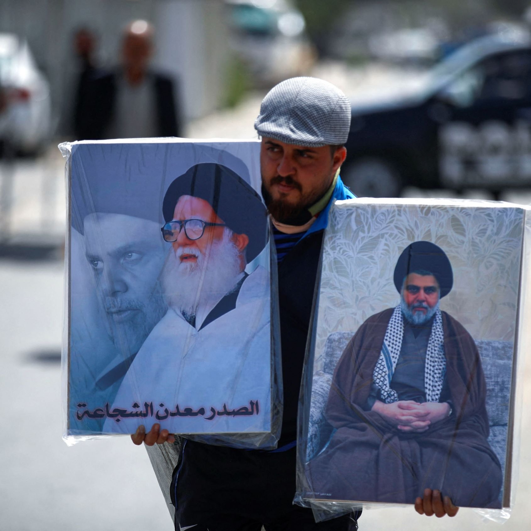 L'immagine mostra un uomo che trasporta due grandi ritratti. A sinistra si vede un'immagine di un leader religioso, mentre a destra c'è un altro ritrato di un diverso leader. L'uomo indossa un berretto e sembra camminare in un contesto urbano, con un veicolo della polizia visibile sullo sfondo. L'atmosfera indica una manifestazione o un evento che riguarda temi religiosi o politici.