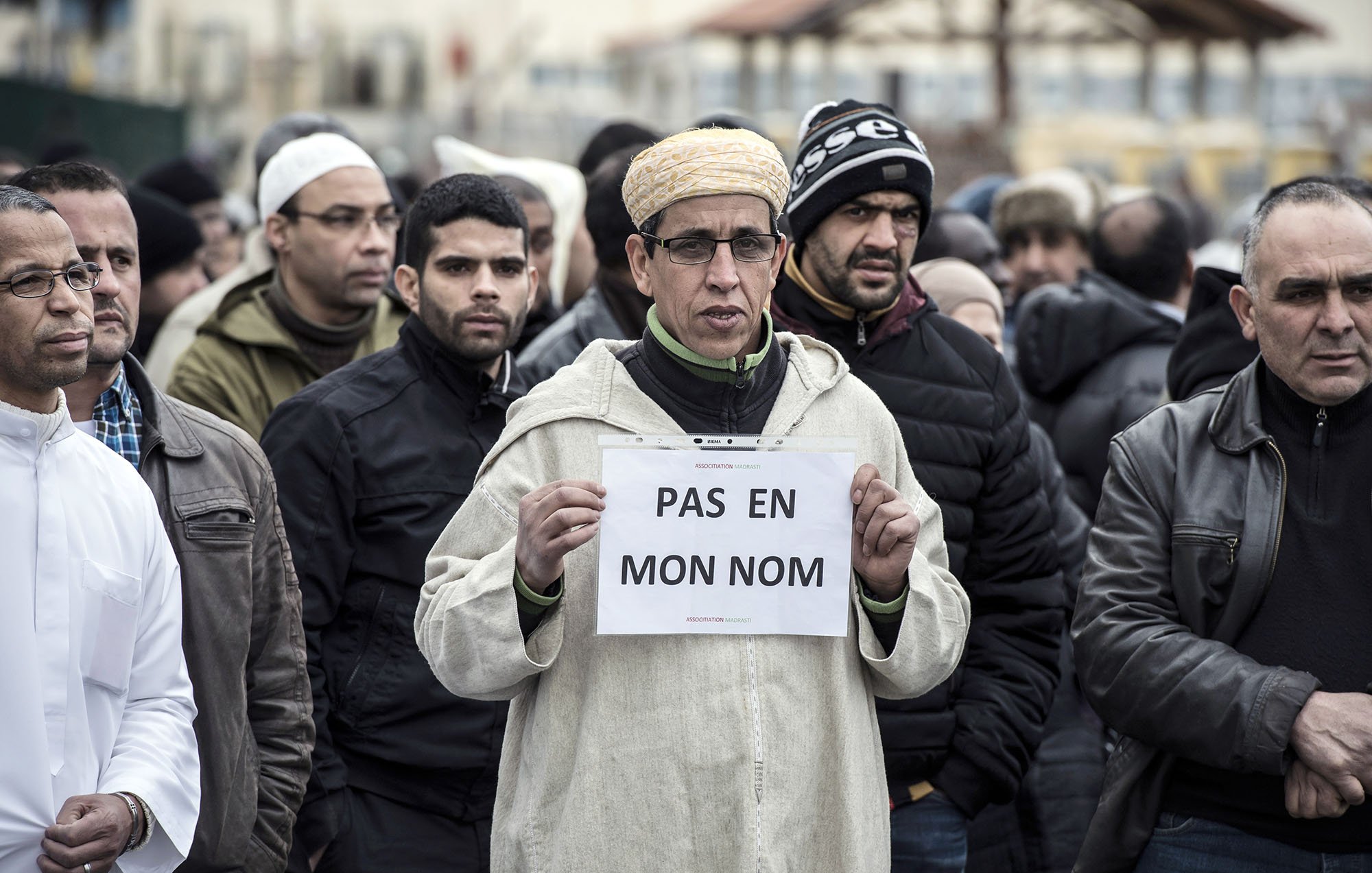 L'image montre un groupe de personnes rassemblées, dont certaines portent des vêtements d'hiver. Au premier plan, un homme se distingue en tenant une pancarte sur laquelle est écrit « PAS EN MON NOM ». Son expression est sérieuse et déterminée. Les autres personnes autour de lui semblent également concentrées sur la situation. L'ambiance générale de la scène suggère une manifestation ou un rassemblement pour une cause particulière.