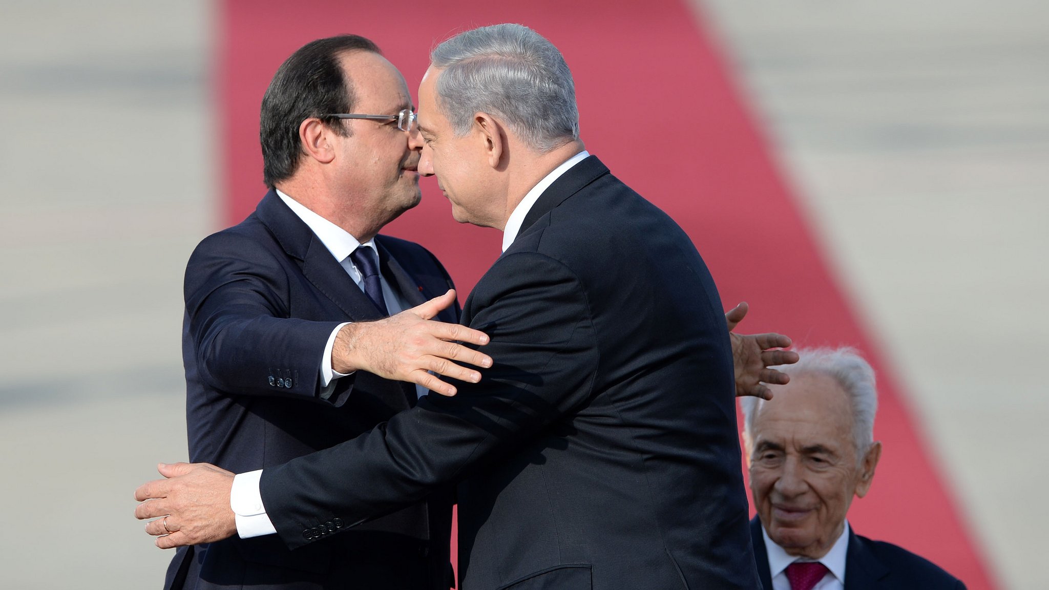 L'image montre deux hommes qui se serrent dans les bras dans un geste amical, tandis qu'un troisième homme est assis derrière eux, regardant la scène. Les deux hommes à l'avant portent des costumes, et on peut notamment voir l'un d'eux pencher la tête en signe de proximité. L'arrière-plan est basé sur une surface rouge, suggérant un événement officiel ou cérémoniel. L'atmosphère semble conviviale et respectueuse.