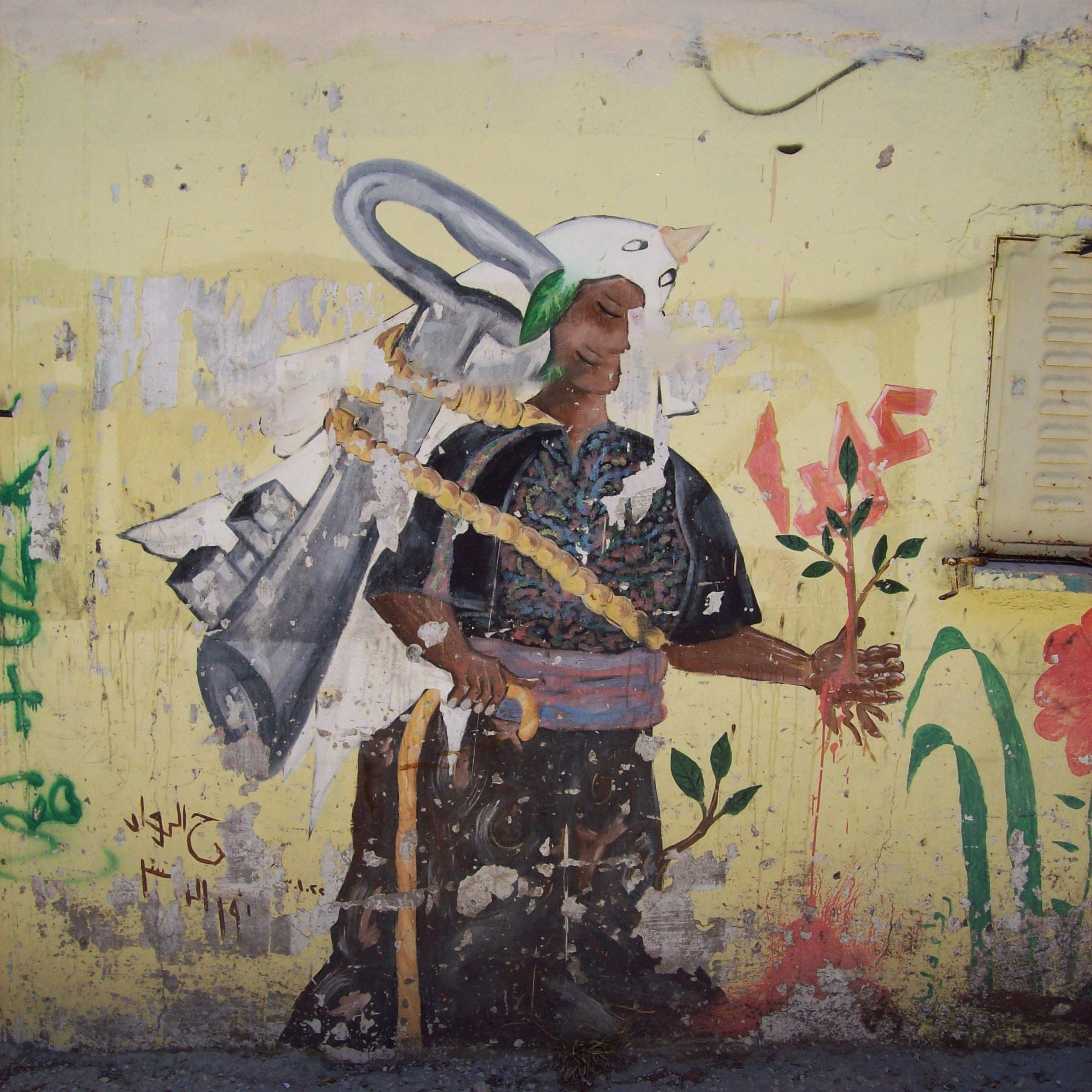 L'image montre une fresque sur un mur. On y voit un personnage portant un costume traditionnel, avec un air déterminé. Ce personnage tient une plante dans une main, symbolisant la vie et la nature. Sur son dos, on peut apercevoir une sorte d'outil ou d'équipement qui pourrait symboliser le travail ou un engagement envers la terre. Le fond est de couleur jaune, ce qui attire l'œil et met en avant le contraste avec les couleurs du personnage. Des inscriptions et des graffitis en arrière-plan ajoutent du caractère à l'œuvre.