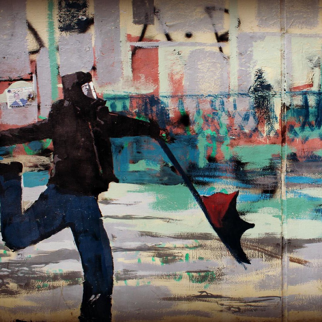 L'image montre une œuvre de street art. À gauche, on voit un homme portant un masque, habillé de noir, qui tient un parapluie ouvert. À droite, une ballerine en robe blanche exécute une danse gracieuse. Le fond de l'œuvre est rempli de couleurs vives et de formes abstraites, créant une atmosphère dynamique. Cette juxtaposition entre les deux personnages évoque un contraste entre l'agitation du monde urbain et la beauté de la danse.