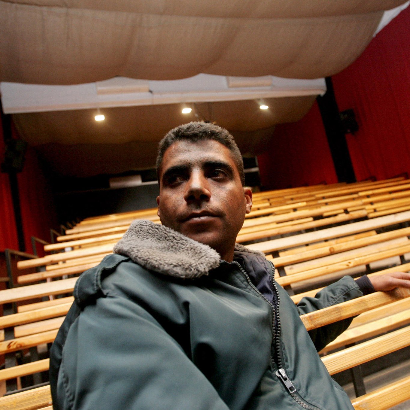 L'image montre un homme assis sur un banc dans un auditorium vide. L'homme porte une veste avec un col en fourrure et semble pensif, regardant vers l'avant. L'arrière-plan est composé de rangées de sièges en bois et de rideaux rouges, créant une atmosphère de théâtre ou de cinéma. La scène est bien éclairée, mettant en valeur l'homme et l'environnement autour de lui.