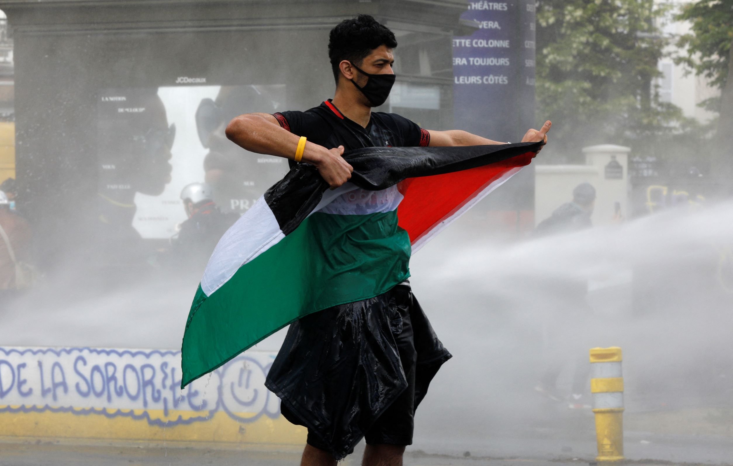 Sur l'image, on voit un jeune homme debout, tenant un drapeau. Il porte un masque noir et semble faire face à une situation de tension, peut-être une manifestation. Le drapeau qu'il brandit a des couleurs verde, blanc et rouge, tandis que de l'eau jaillit autour de lui, suggérant l'utilisation de canons à eau. L'environnement semble être urbain, avec des signes de protestation en arrière-plan.