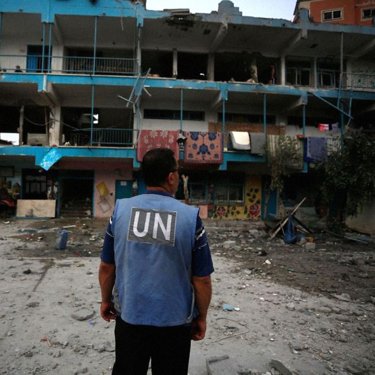 The image depicts a scene of destruction, likely from a conflict area. In the foreground, a person wearing a blue vest with "UN" printed on it stands, facing a damaged building with shattered windows and debris scattered around. The building has signs of neglect, with graffiti and remnants of what may have been a children's play area. People can be seen in the background, possibly assessing the situation or providing assistance. The overall atmosphere conveys a sense of urgency and the aftermath of turmoil.