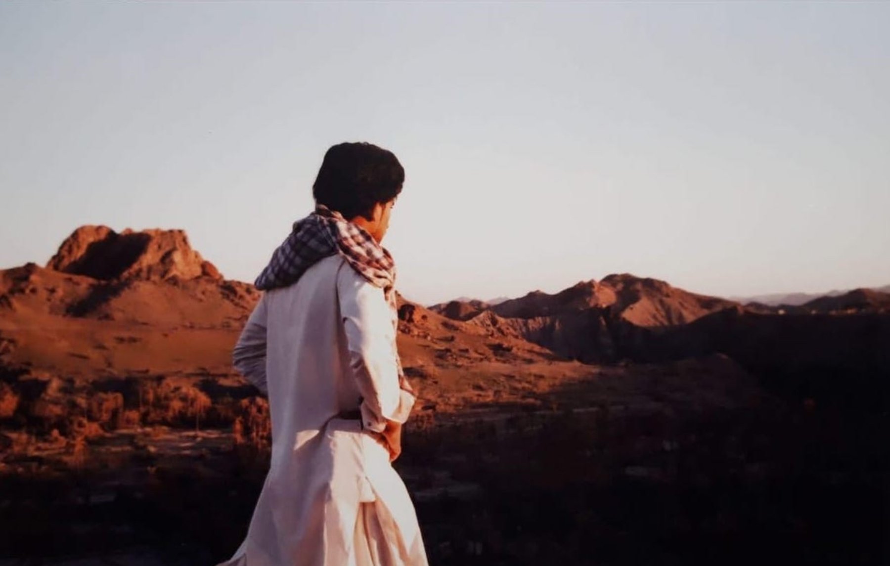 L'image montre une personne debout sur une colline, regardant au loin vers un paysage montagneux. La lumière douce du coucher de soleil illumine les montagnes environnantes, créant une atmosphère sereine. La personne porte une tenue traditionnelle, avec un chèche sur la tête, qui évoque une culture spécifique. L'arrière-plan est composé de collines et de paysages arides, typiques de certaines régions désertiques.