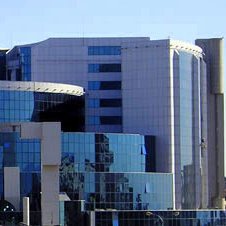 L'image montre un bâtiment moderne au design architectural contemporain. La façade est principalement en verre, ce qui reflète la lumière et les bâtiments environnants. Des formes géométriques et des courbes sont bien mises en valeur, et on peut distinguer des éléments en métal. Le paysage autour est agrémenté d'arbres et de plantes, suggérant que le bâtiment est situé dans une zone urbaine. L'ensemble dégage une impression de sophistication et de modernité.