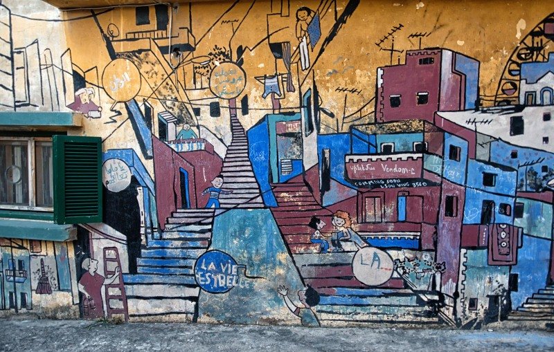 L'image montre une murale colorée peinte sur un mur, représentant une scène de la vie urbaine. On y voit des escaliers, des maisons aux façades vives, et des personnages en train de vaquer à diverses activités. La murale semble évoquer la culture et l'esprit d'une communauté, avec des mots tels que "LA VIE" et "ESBELLE" intégrés dans le dessin. Les couleurs utilisées sont vives et variées, ajoutant une ambiance dynamique et joyeuse à la scène.