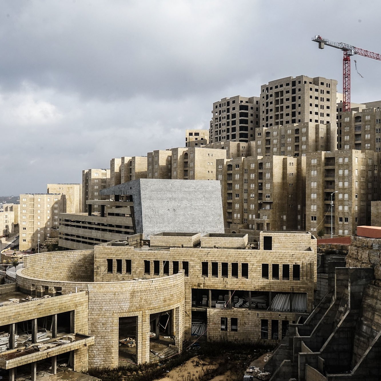 L'image montre un paysage urbain en construction, avec des bâtiments modernes de forme géométrique et des structures encore inachevées. On peut voir des façades en béton et des fenêtres non terminées sur plusieurs des bâtiments. En arrière-plan, une grue rouge indique que des travaux sont en cours. Le ciel est nuageux, créant une atmosphère un peu sombre. Au premier plan, des éléments de construction sont visibles, suggestifs d'un site en développement.