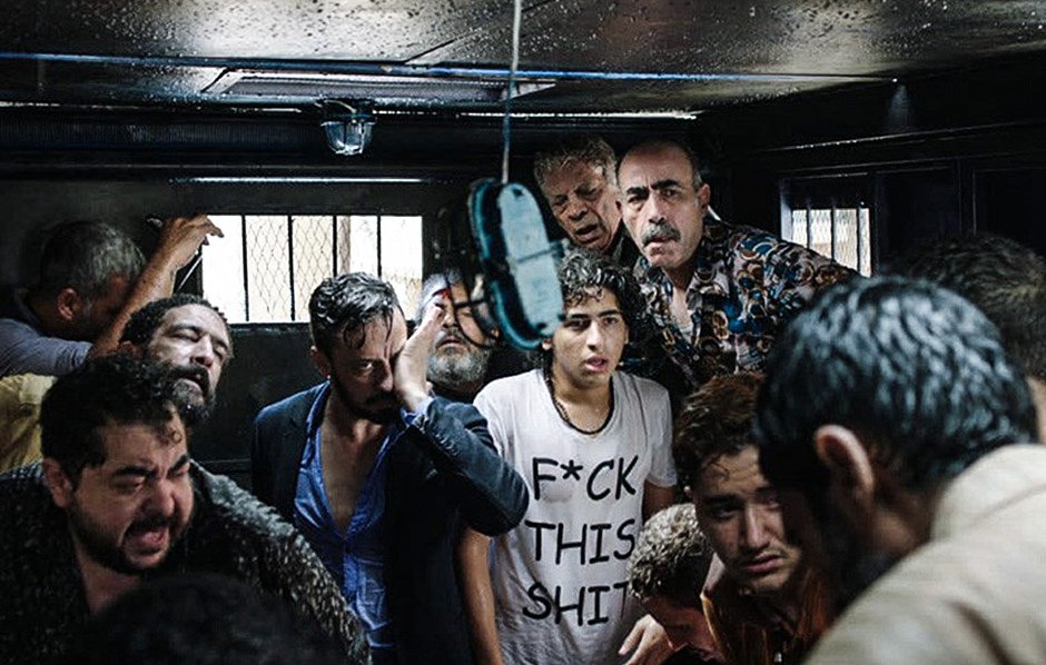 L'image montre un groupe de personnes dans un espace clos, visiblement en détresse ou en désespoir. Les visages expriment des émotions fortes, allant de la peur à l'angoisse. Certains sont habillés de manière décontractée, et l'un des individus porte un t-shirt avec un message provocateur. L'éclairage est faible, créant une ambiance sombre et oppressante. L'environnement suggère une situation de tension ou de crise.