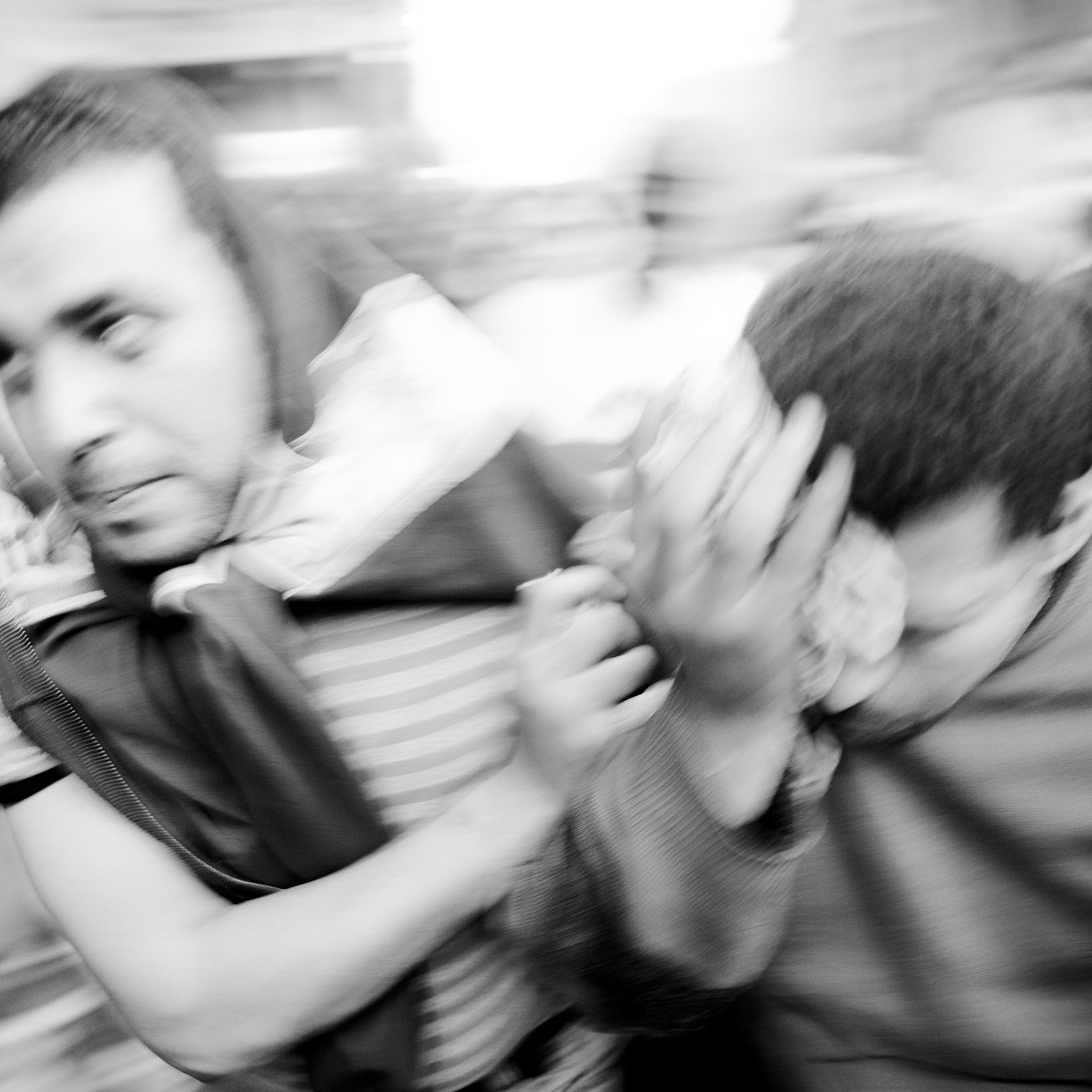 L'image montre deux personnes en mouvement dans un environnement flou, ce qui donne une impression de dynamisme et d'urgence. L'une des personnes semble porter quelque chose sur son épaule, tandis que l'autre se couvre le visage avec une main. L'atmosphère est intense, comme si les sujets étaient en train de fuir ou de réagir à une situation pressante. Les couleurs sont principalement en noir et blanc, accentuant le contraste et l'émotion de la scène.