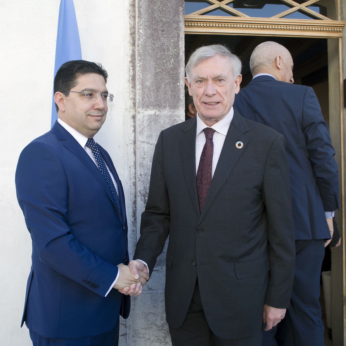 L'image montre deux hommes en train de se serrer la main, échangeant probablement une salutation ou un accord. L'un est habillé d'un costume bleu, tandis que l'autre porte un costume sombre avec une cravate. On aperçoit un drapeau derrière eux, suggérant un contexte officiel ou diplomatique. Leur expression semble amicale et professionnelle.