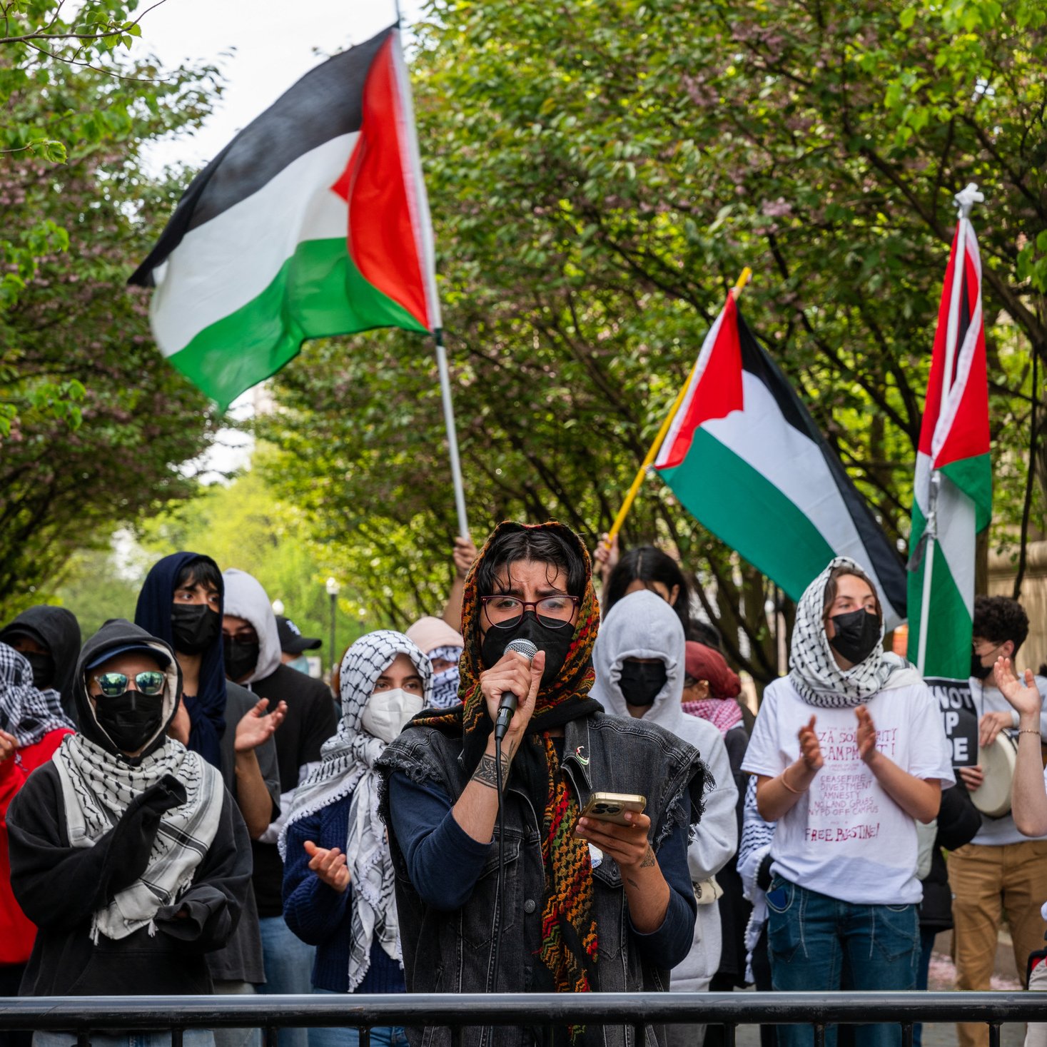 L'image montre une manifestation en plein air où plusieurs personnes sont rassemblées. Au premier plan, une personne prend la parole avec un microphone, vêtue de vêtements sombres et entourée de manifestants portant des masques. Beaucoup des présents arbore des foulards ou keffiehs. En arrière-plan, des drapeaux palestiniens flottent, ajoutant de la couleur à la scène. Les manifestants semblent soutenir une cause, probablement liée à la Palestine, et il y a une atmosphère de solidarité parmi le groupe. Les arbres en fleurs encadrent la scène, créant un contraste avec l'intensité de la manifestation.