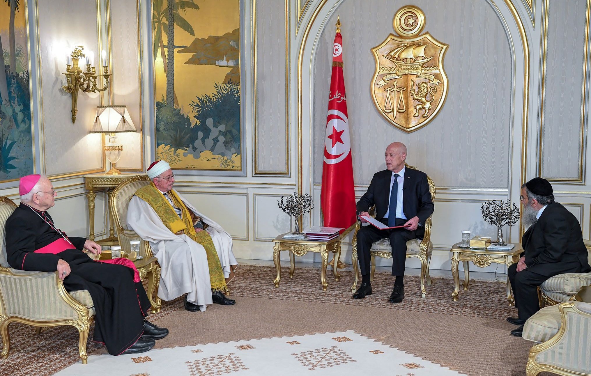 L'image montre une réunion dans un cadre formel. On peut voir plusieurs personnes assises autour d'une table basse. À gauche, un homme en soutane rouge, probablement un évêque, et un homme en robes traditionnelles, au centre, qui semble être un chef religieux. À droite, un homme en costume noir. Au fond, le drapeau tunisien flotte, et on peut apercevoir des éléments de décoration élégants sur les murs et des plantes en arrière-plan. La scène évoque un moment de dialogue interreligieux ou de rencontre officielle.