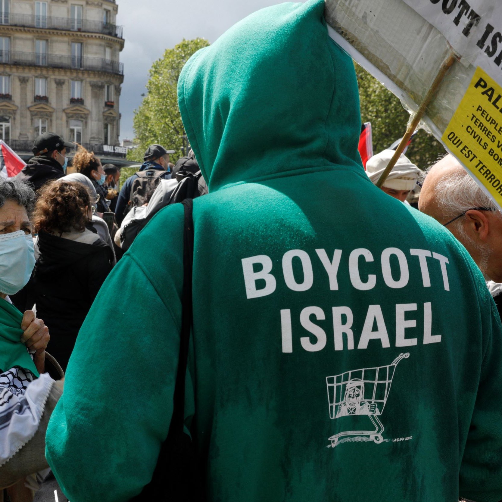 تظهر في الصورة مجموعة من الأشخاص في تظاهرة تتعلق بقضية فلسطين. يرتدي أحد المتظاهرين سترة خضراء مكتوب عليها "BOYCOTT ISRAEL"، مما يعبر عن دعوة لمقاطعة إسرائيل. في الصورة أيضًا، هناك امرأة ترتدي كمامة وتبدو أنها تتحدث مع المتظاهرين الآخرين. الخلفية تشمل مباني ورايات تحمل شعارات مختلفة. الجو العام يوحي بالتضامن والمشاركة في حركة مناهضة للاحتلال.