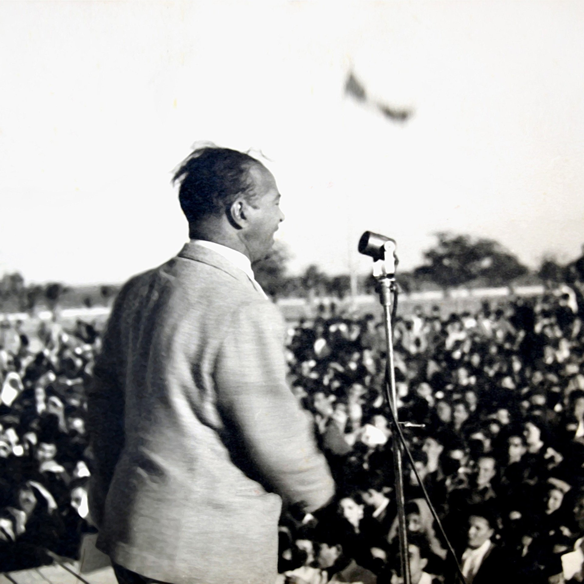 L'image montre un homme en train de parler sur une scène, devant un grand public. Il est vu de dos, et on peut voir une foule nombreuse devant lui, captivée par son discours. On distingue également un microphone sur le podium. L'atmosphère semble dynamique et engagée, probablement lors d'un événement politique ou d'un rassemblement. En arrière-plan, il y a des drapeaux, ce qui pourrait indiquer un contexte symbolique ou patriotique.