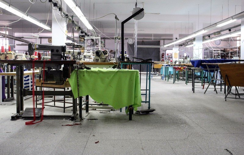 L'image montre un atelier de couture ou de fabrication. On y voit plusieurs tables de travail avec des machines à coudre. Certaines machines semblent être à l'arrêt, et des morceaux de tissu sont éparpillés sur le sol. L'éclairage est assez lumineux, et l'espace semble vaste mais légèrement en désordre, ce qui donne une impression d'activité récente. Des outils et du matériel de couture sont visibles sur les surfaces de travail, et le décor est fonctionnel et utilitaire.