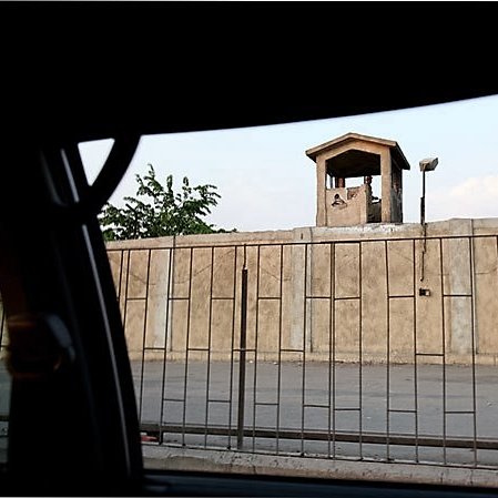 تظهر في الصورة جزء من سياج عازل يحيط بمكان ما، مع وجود برج مراقبة صغير في الأعلى. يمثل البرج نقطة مراقبة، بينما الأسفل يظهر حاجزًا يفصل بين الشارع والفضاء المحيط. تضيء السماء بلونٍ فاتح، مما يدل على أن الوقت قد يكون في فترة المساء أو الصباح. يبدو أن الصورة قد تم التقاطها من داخل مركبة، حيث يظهر إطار السيارة من الجهة اليسرى.