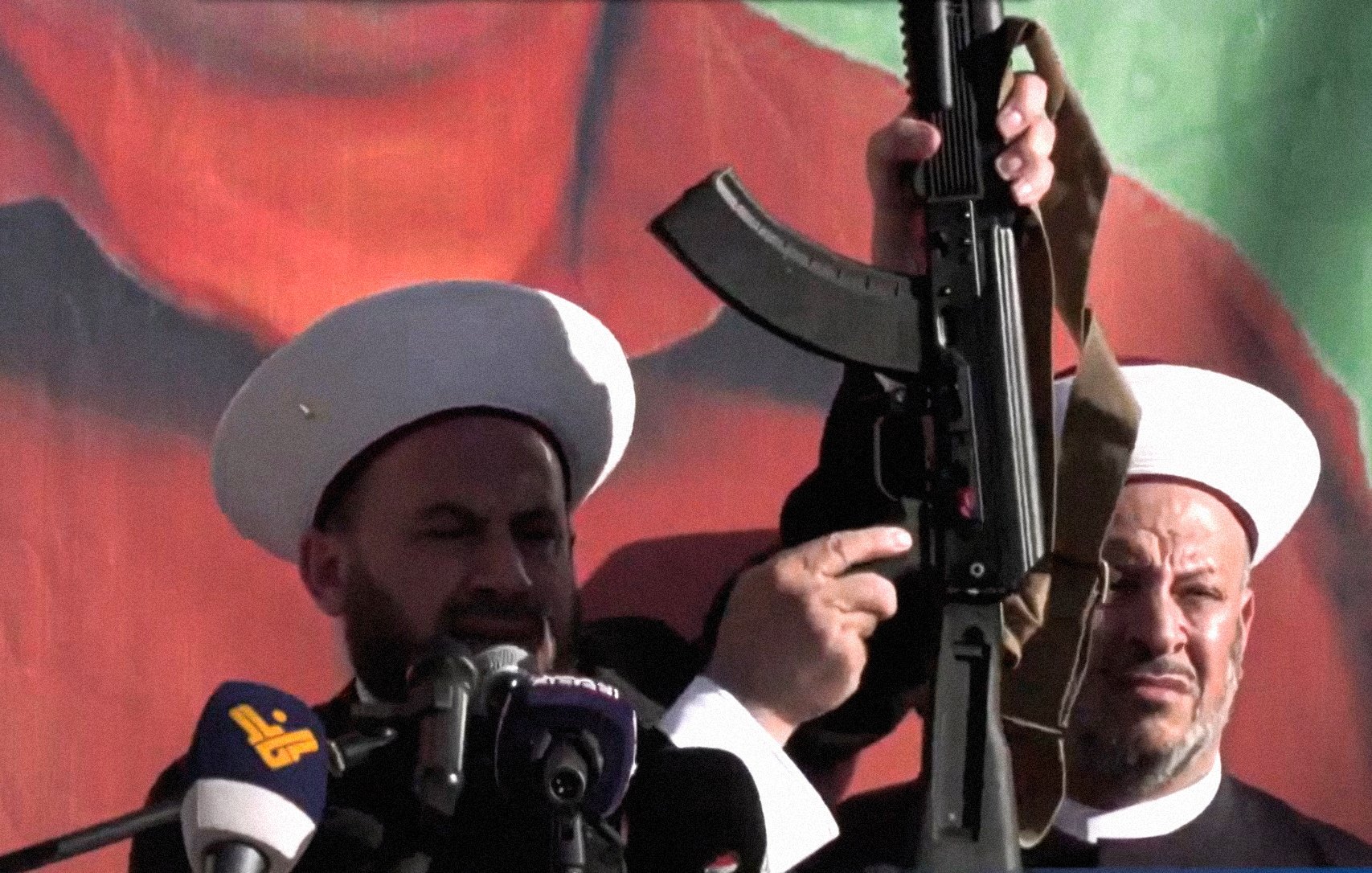 L'immagine mostra due uomini in abiti tradizionali, entrambi con copricapi bianchi, che sembrano partecipare a un evento pubblico o a una manifestazione. Uno di loro tiene in mano un fucile d'assalto, mentre l'altro osserva. Sullo sfondo c'è un grande murale o una bandiera che potrebbe rappresentare simboli politici o culturali. L'atmosfera sembra intensa e carica di emozione, con un focus sulla figura che tiene l'arma.