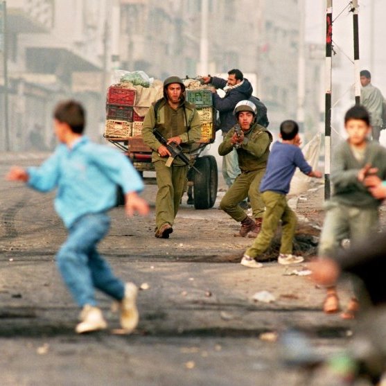 L'image montre une scène de tension dans un environnement urbain, où deux soldats sont présents, armés et en position d'action. Un homme, probablement un civil, est également visible, tandis que des enfants et d'autres personnes s'éloignent rapidement de la situation, suggérant un climat de peur ou de chaos. Des véhicules et des baraques sont en arrière-plan, accentuant l'atmosphère de conflit. L'ensemble évoque un contexte de violences ou d'affrontements dans une ville.