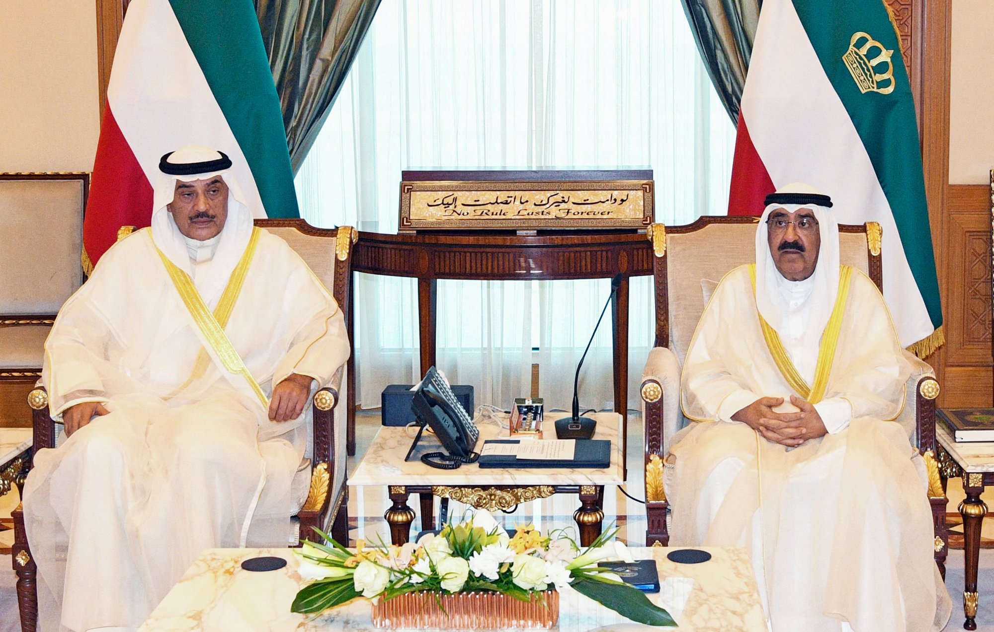 L'image montre deux hommes assis dans une salle de réunion élégante. Ils portent des vêtements traditionnels, possiblement des habitudes de type arabe, avec des écharpes sur la tête. Leurs expressions sont sérieuses et ils sont entourés de décorations luxueuses, comme des rideaux drapés et des meubles en bois. Au centre, il y a une table en marbre avec quelques objets, et en arrière-plan, on peut voir un tableau avec des inscriptions arabes. L'atmosphère dégage un sentiment de formalité et de respect.
