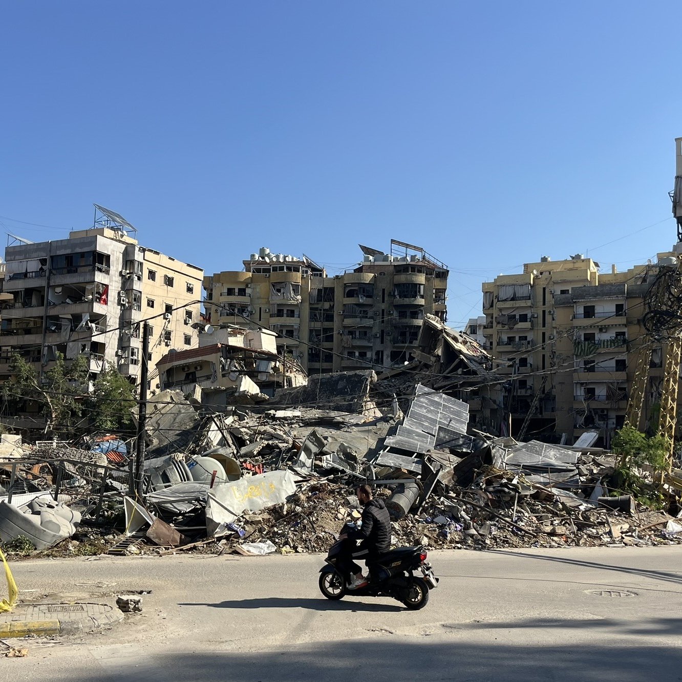 L'image montre une scène de destruction urbaine, où plusieurs bâtiments sont partiellement effondrés et en ruines. On peut voir des décombres éparpillés au sol, tandis qu'une personne à moto passe devant. Le ciel est dégagé, laissant transparaître un jour ensoleillé, contrastant avec le désastre visible. La scène évoque un fort sentiment de désolation et de dégâts, typique de zones touchées par des conflits ou des catastrophes.