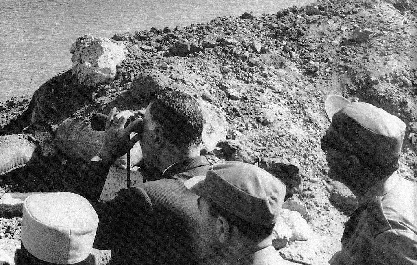 L'image montre un groupe d'hommes, dont l'un semble observer quelque chose au loin à l'aide de jumelles. Ils se tiennent près d'une rive d'eau, probablement à proximité d'un site de construction ou d'une digue, entourés de débris ou de terre. L'environnement semble suggérer une période historique, peut-être liée à des événements militaires ou politiques. Les hommes portent des uniformes ou des vêtements formels, ce qui indique qu'ils occupent probablement des positions officielles.