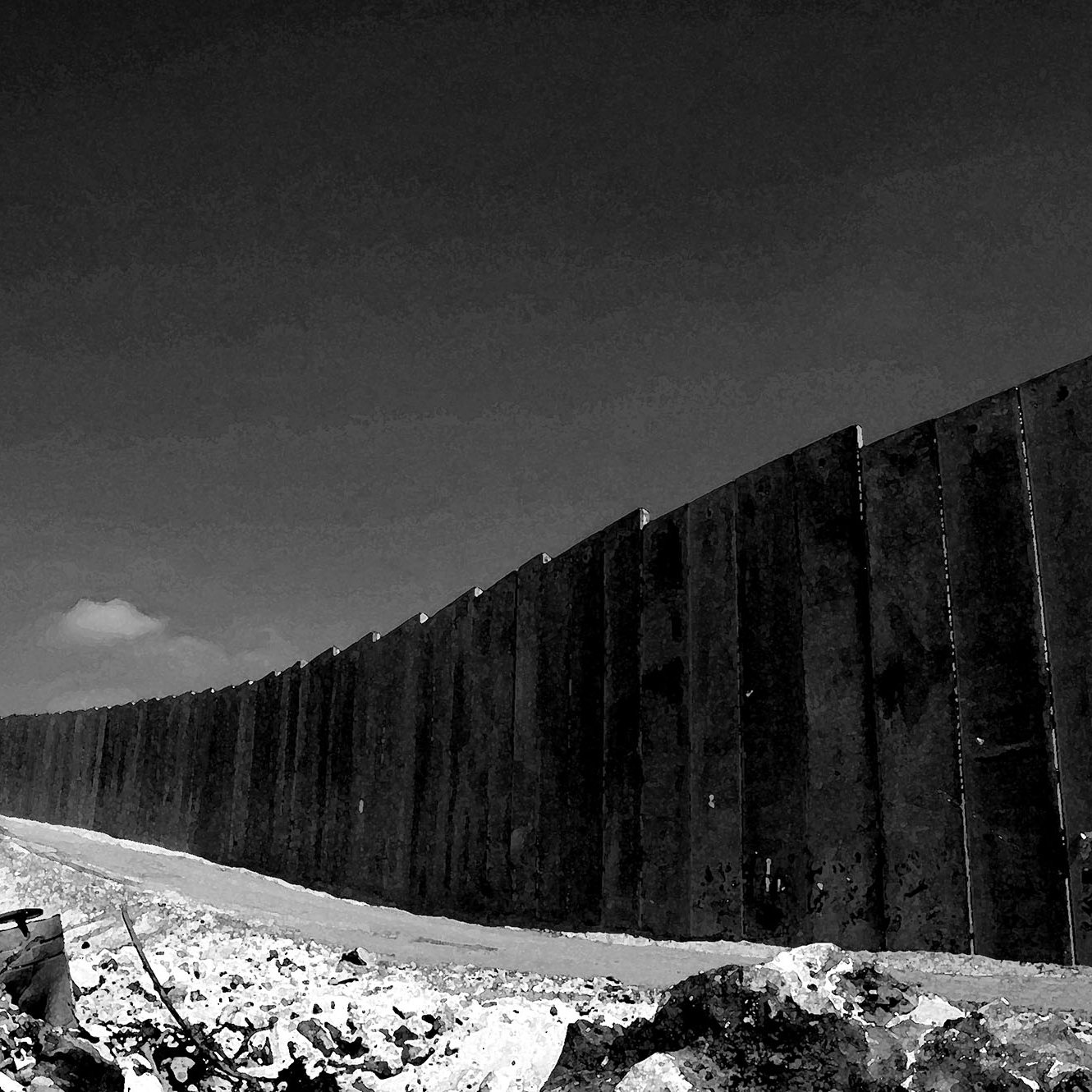 L'immagine mostra un muro lungo e imponente, eretto in un paesaggio desolato. La scena è in bianco e nero, il che le conferisce un'atmosfera drammatica. Il muro, costituito da grandi pannelli di cemento, si estende in modo curvilineo verso l'orizzonte, mentre il terreno attorno appare roccioso e poco curato. In lontananza, si intravede un cielo chiaro, con una nube che si staglia sullo sfondo, aggiungendo profondità all'immagine. L'assenza di colori enfatizza la severità e la rigidità della struttura, suggerendo un senso di divisione e separazione.