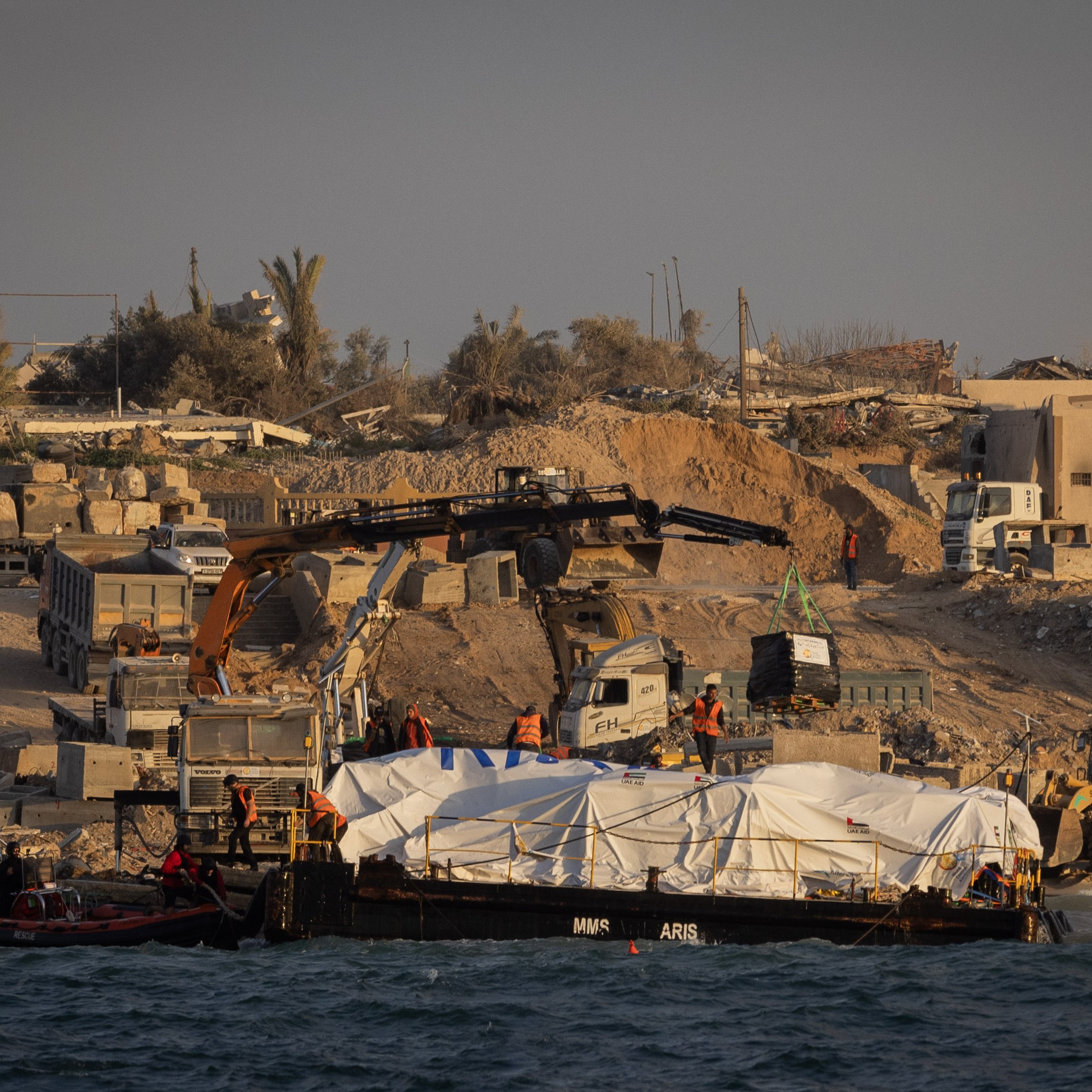 L'image montre une scène de construction ou de travaux sur une côte. On peut voir un bateau sur l'eau avec une grande bâche blanche, probablement transportant des matériaux ou des fournitures. Sur la rive, il y a des machines de construction, comme des pelleteuses et des camions, ainsi que des travailleurs en action. En arrière-plan, des bâtiments ruinés et des palmiers suggèrent un environnement en mutation, peut-être en raison de travaux de développement ou de reconstruction. L'atmosphère générale semble celle d'un chantier en cours.