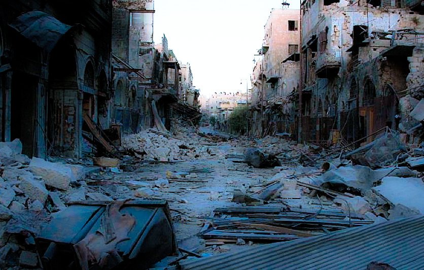 L'image montre une rue dévastée, probablement après un conflit ou une catastrophe. On peut voir des bâtiments endommagés avec des murs effondrés et des débris éparpillés sur le sol. Le contexte semble être celui d'une ville abandonnée, avec une atmosphère sombre et désolée. Les traces de destruction sont évidentes, illustrant les ravages subis par l'environnement urbain.